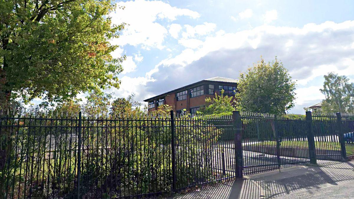 A school building set against bright skies is behind high black metal fencing. There are bushes and trees in the grounds of the school.