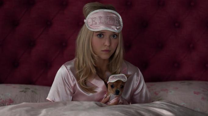 A woman with long blonde hair sits up in bed, with pink bedsheets, pink satin PJs and a pink eye mask on her forehead that has "ELLE" written in diamontes. She's holding a tiny dog which also has a little eye mask on.