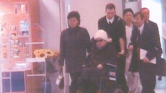 Ko Yong Hui, in sunglasses and a white beanie, sits in a wheelchair pushed by a man. Several people are around her at a hospital in France