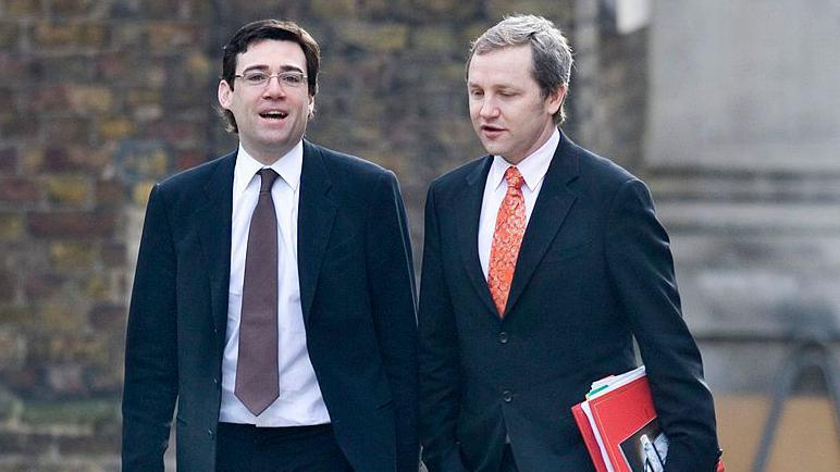 Andy Burnham and James Purnell walking down Downing Street. Both men are wearing dark-coloured suits, white shirts and ties. Purnell is holding a red-coloured folder.