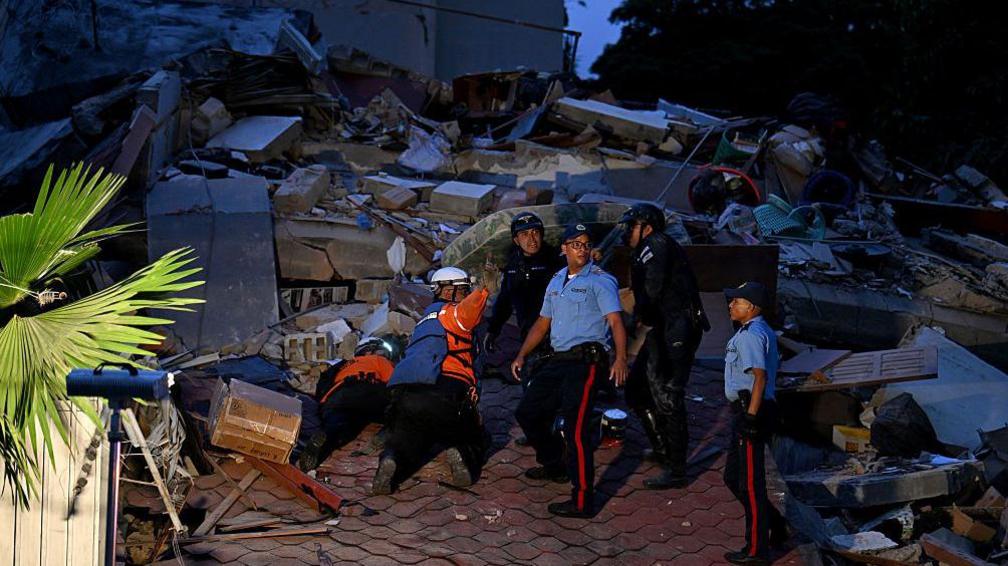 An image of six people standing on a pile of rubble, searching for people. They are wearing emergency worker uniforms and four have safety helmets on. The sky is dark.