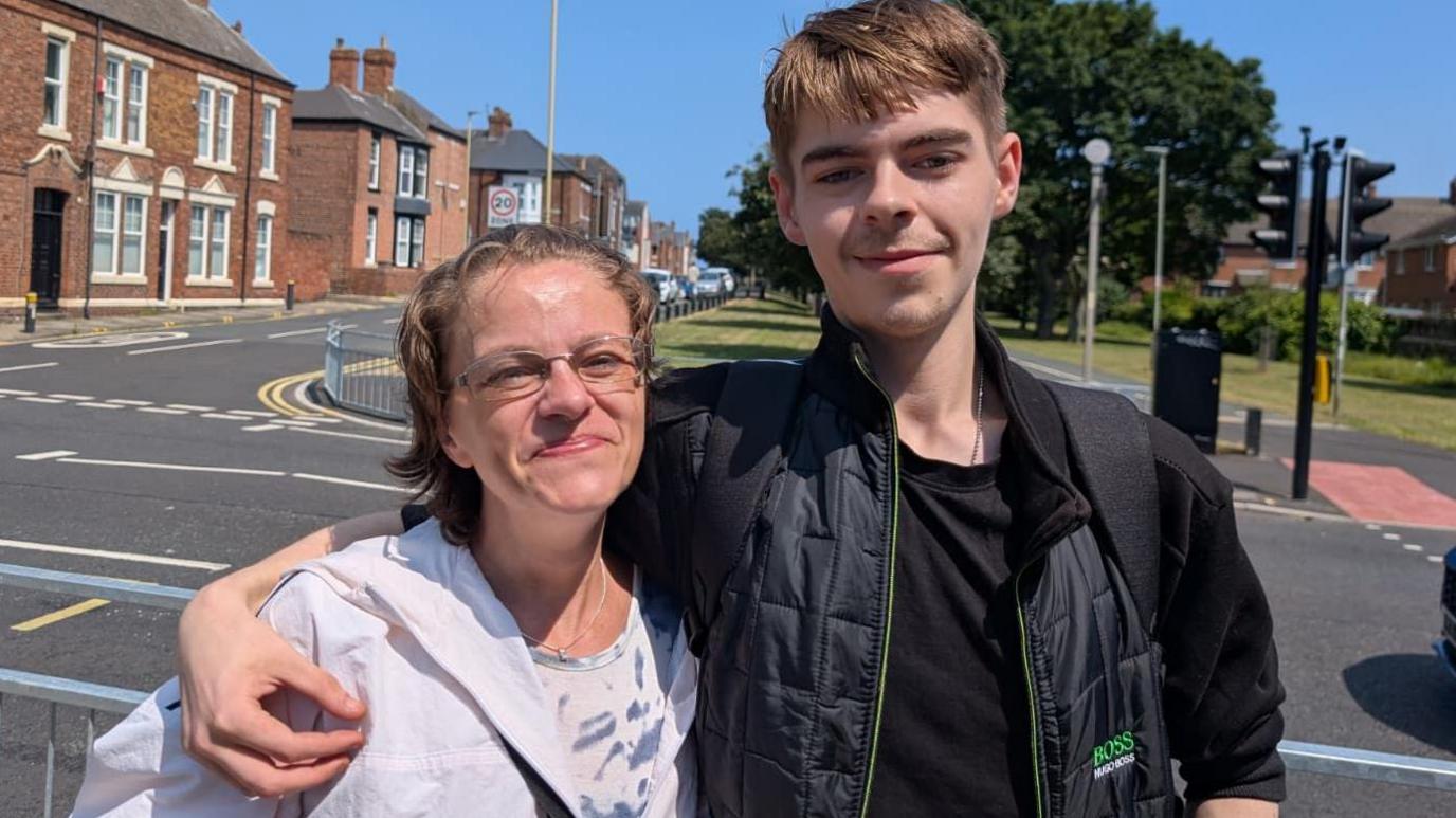 Nic, a woman in her 40s with short swept back hair and rectangular glasses smiles warmly next to her son Scott who has his arm around her. He's about a foot taller than her and has a regular brown hair cut. He's wearing a black Hugo Boss bomber jacket while Nic wears a lilac and white tie abstract patterned shirt and a light cagoule.
