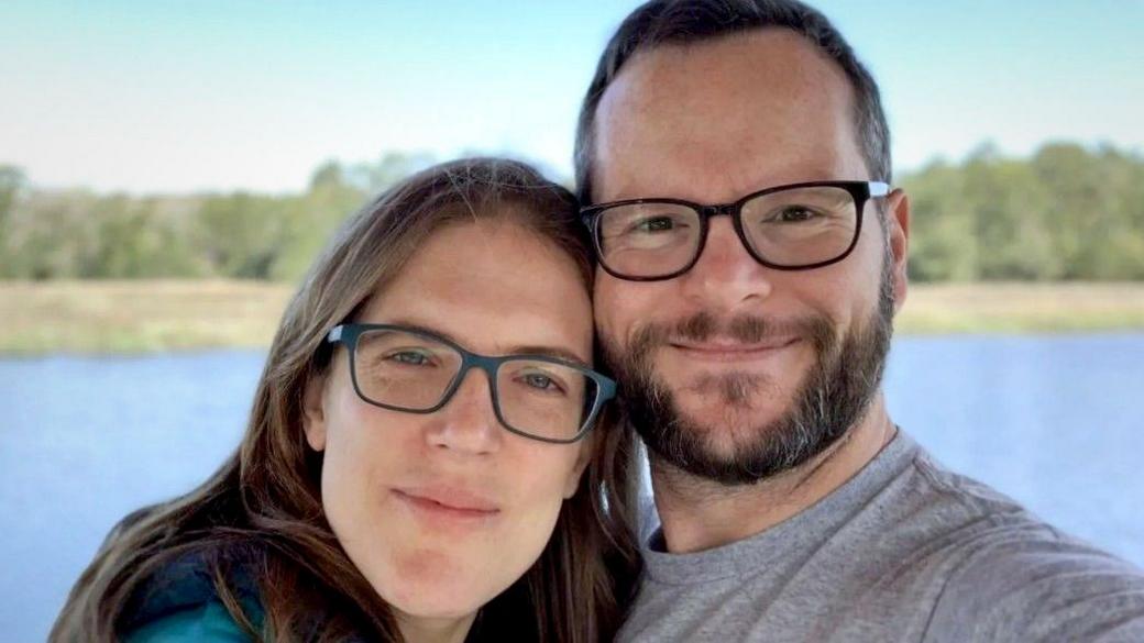 Katie and Alan Donegan smile at the camera while both wearing glasses during a selfie in front of a lake and trees against a blue sky.