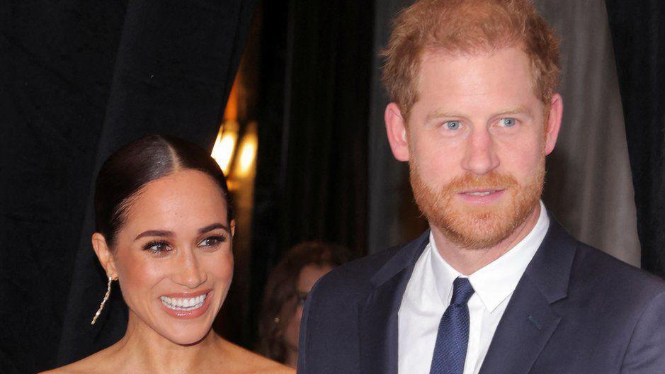 A smiling Meghan Markle stands next to Prince Harry, who wears a blue suit and tie with a white shirt.  She wears long gold-coloured earrings.