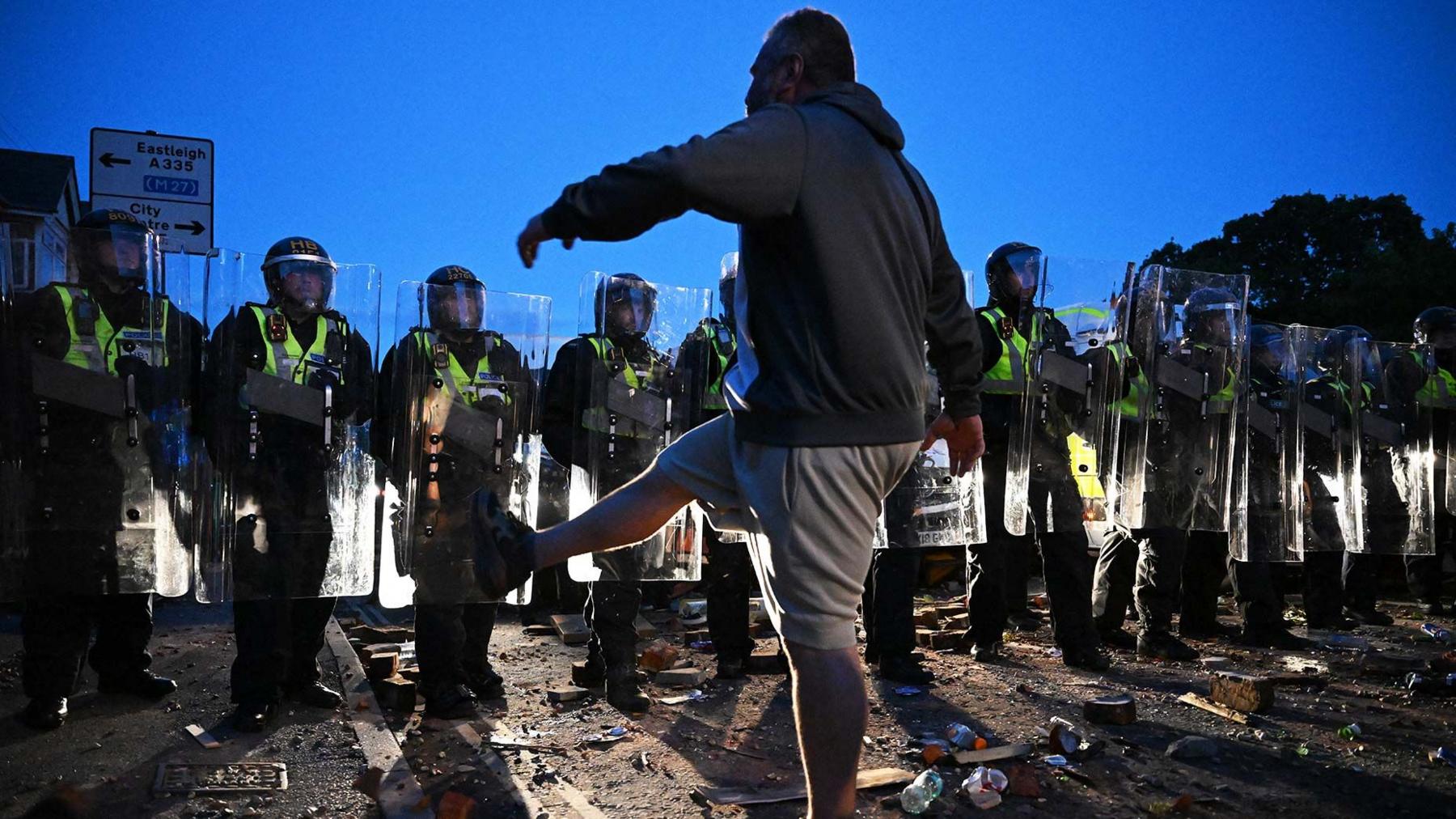 Police in Southampton line up in a clash against protesters . A male protester is seen in the foreground  silhouetted against the sky kicking his leg at the police 