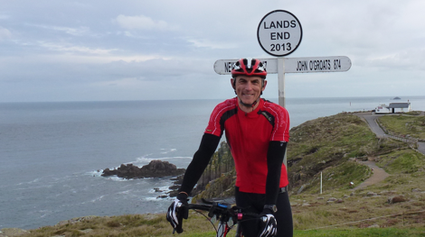 A man is wearing a red cycling top and black lyrca while holding a bike next to the sea and a sign for Lands End