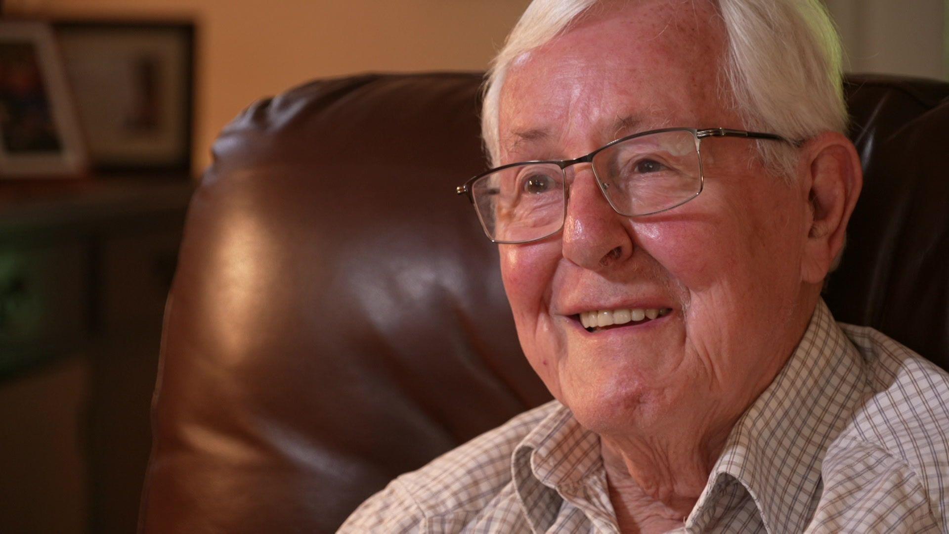 Stephen Harrington is an older man, aged 83. He has white hair and wears thin-rimmed clear-lensed glasses. He is sitting on a brown leather chair and wears a white and brown checked shirt. He is smiling.