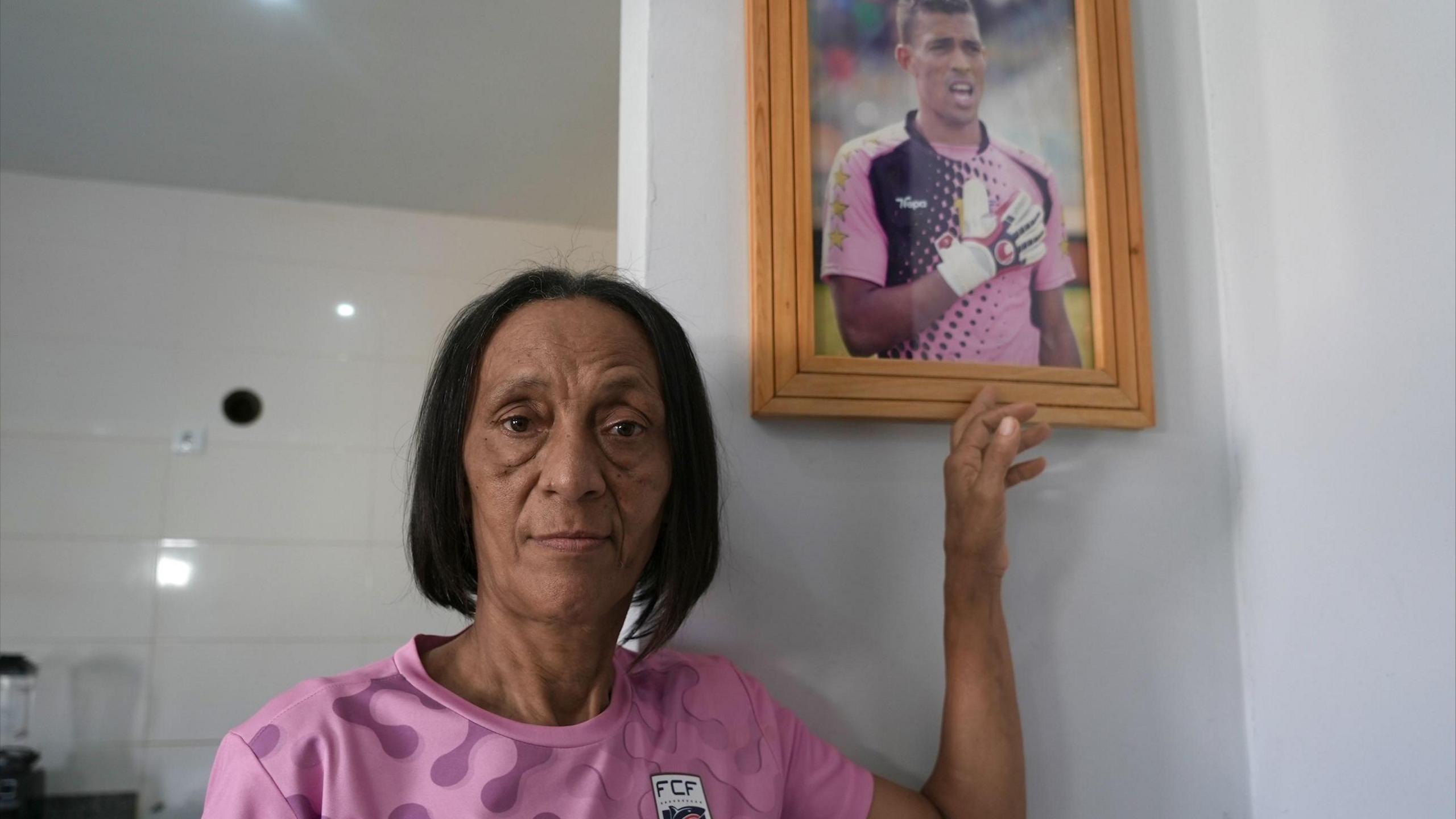 Vozinha's mother stands next to a framed photo of her son at her home.