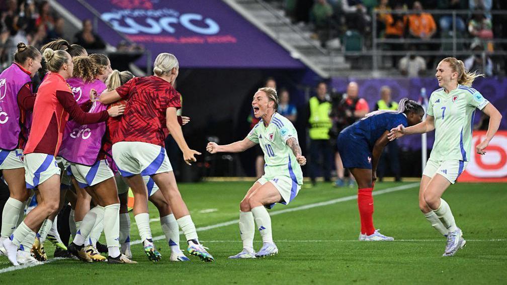 Jess Fishlock in a green Wales change shirt celebrates as she approaches subsitutes gathered on the touchline at the Wales v France game in Euro 2025. Team-mate Ceri Holland is running towards them.