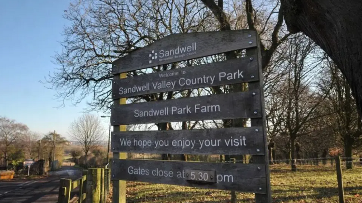 A mainly dark sign that includes the words Welcome to Sandwell Valley Country Park. Trees and grass are in the background.