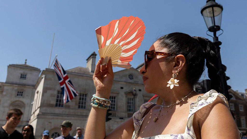 A woman wearing a white dress and tortoiseshell sunglasses puts an orange fan above her head in central London