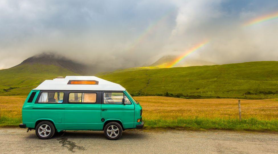 A traditional-style VW campervan parked on a road next to fields and mountains. They are slightly obscured by low cloud, but a rainbow is also visible in the background.