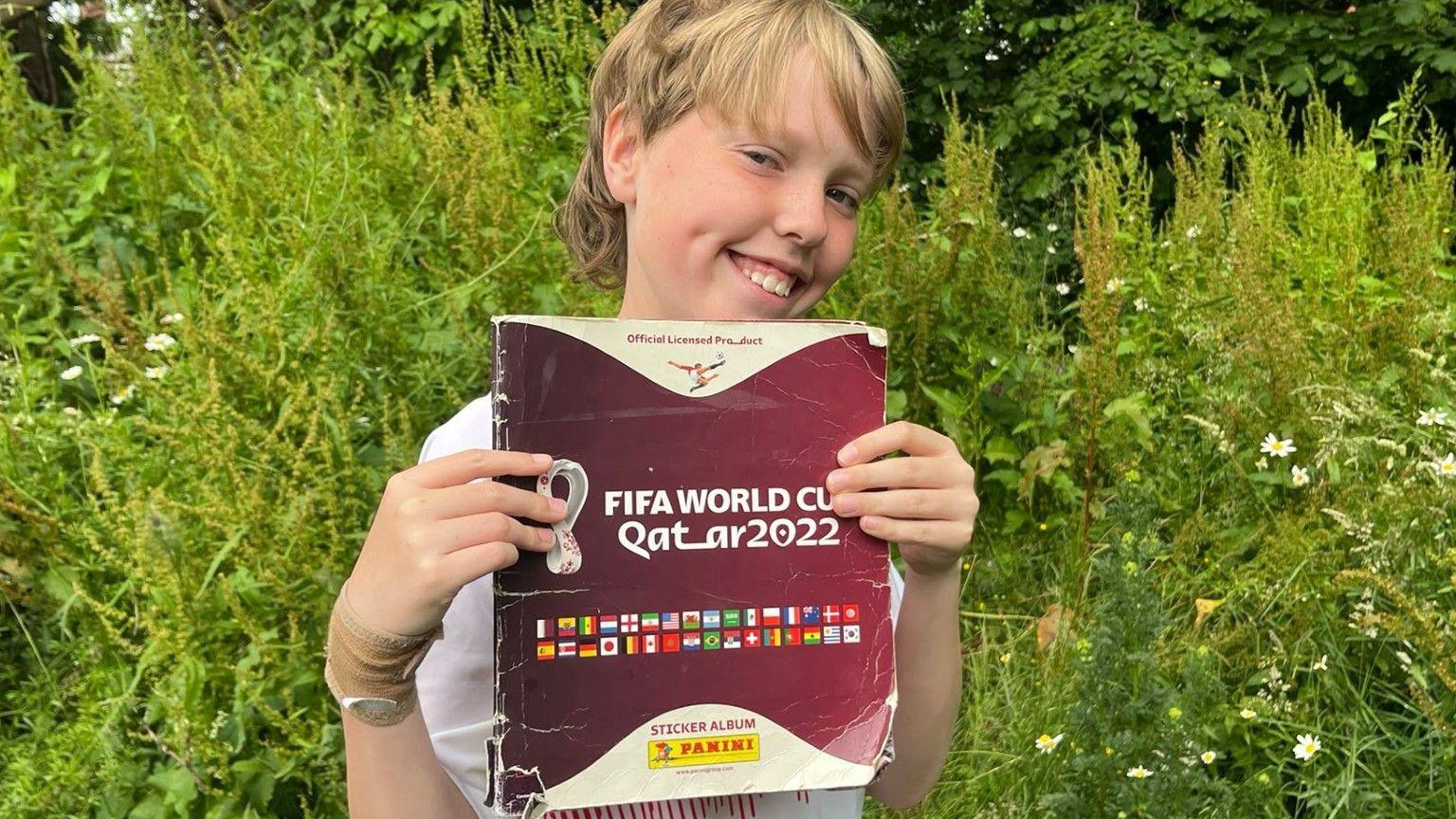 A boy with longer blonde hair stands in front of some trees. He is smiling. He is holding up the 2022 Panini Qatar World Cup sticker book, which has a maroon cover with the flags of the competing countries, and the logo of the tournament. It is slightly crumpled and creased.