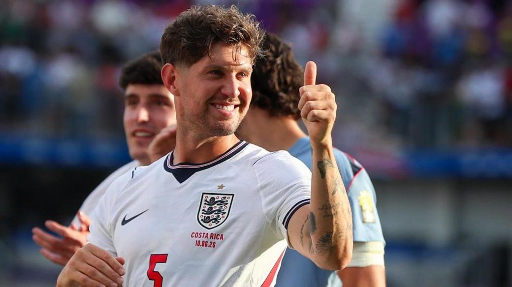 John Stones gives a thumbs-up gesture while he wears white England kit that includes wording 'Costa Rica 10.06.26' below the Three Lions badge