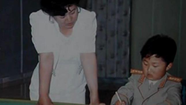 Ko Yong Hui, in a white dress, stands on the left. A young Kim Jong Un, in a miitary uniform, sits and writes with a pen.