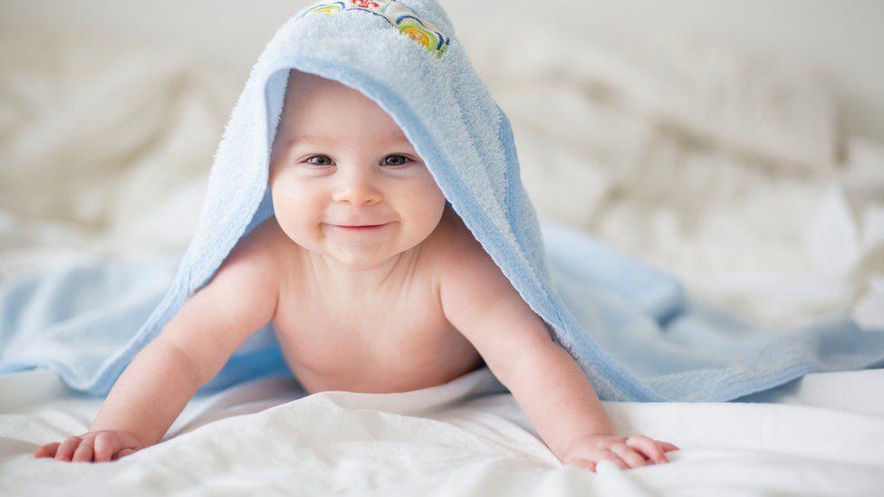 A smiling baby lies on a bed wearing a light blue hooded towel.