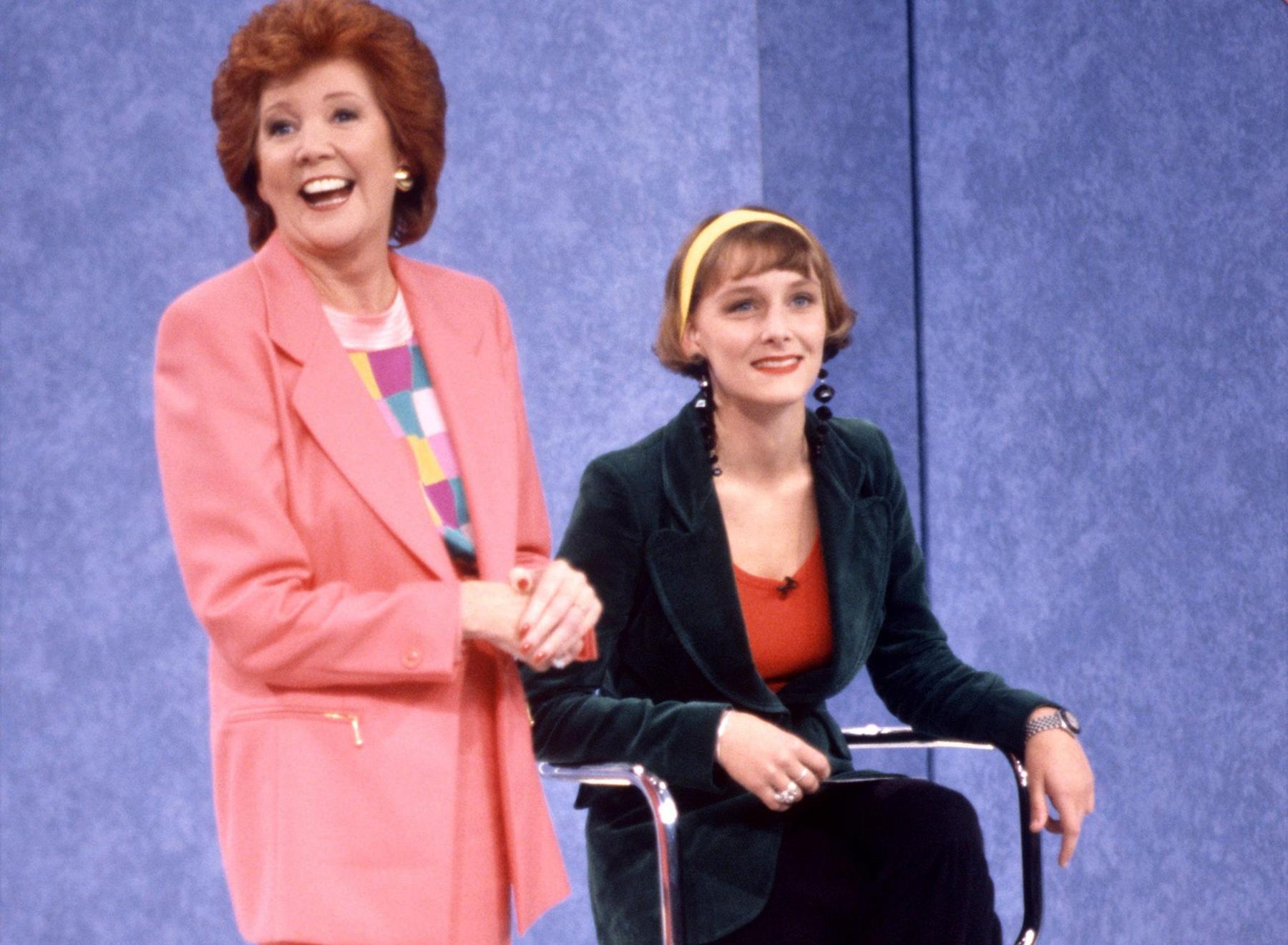 Frankie (R) is smiling and sitting on a chair next to Cilla Black, who is standing. Frankie is wearing a bottle green blazer over an orange-red top. Her blonde hair is styled in a short bob with a fringe and held back by a bright yellow hairband. Cilla is wearing a peach jacket over a checked, pastel top, with red nail varnish. Her signature auburn hair is worn in a shoulder-length bouffant style.