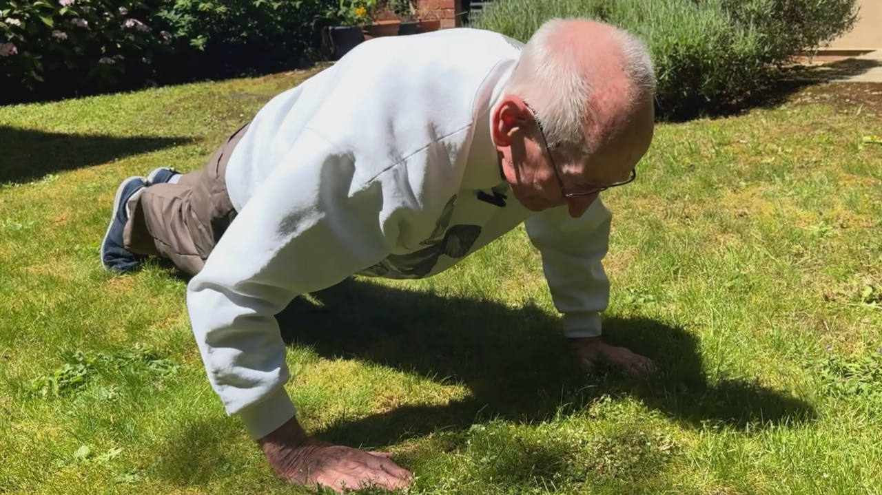 An elderly man doing a press-up on grass in a garden.