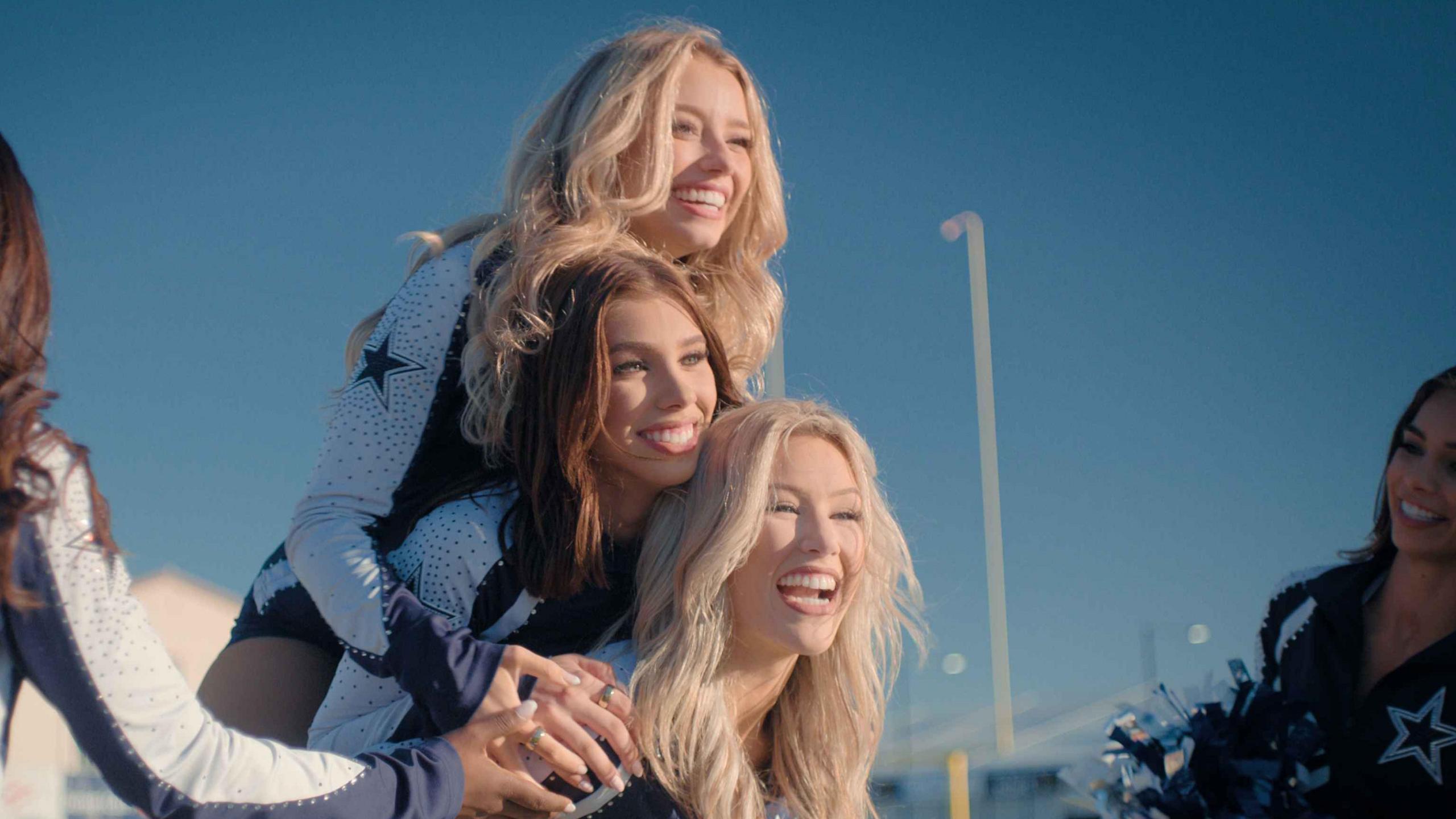 Pictured from  top to bottom: Kleine, Lea, Megan. Kleine and Megan have long blonde hair and Lea has long brunette hair. They are shown jumping on each other while two squad members stand at the side