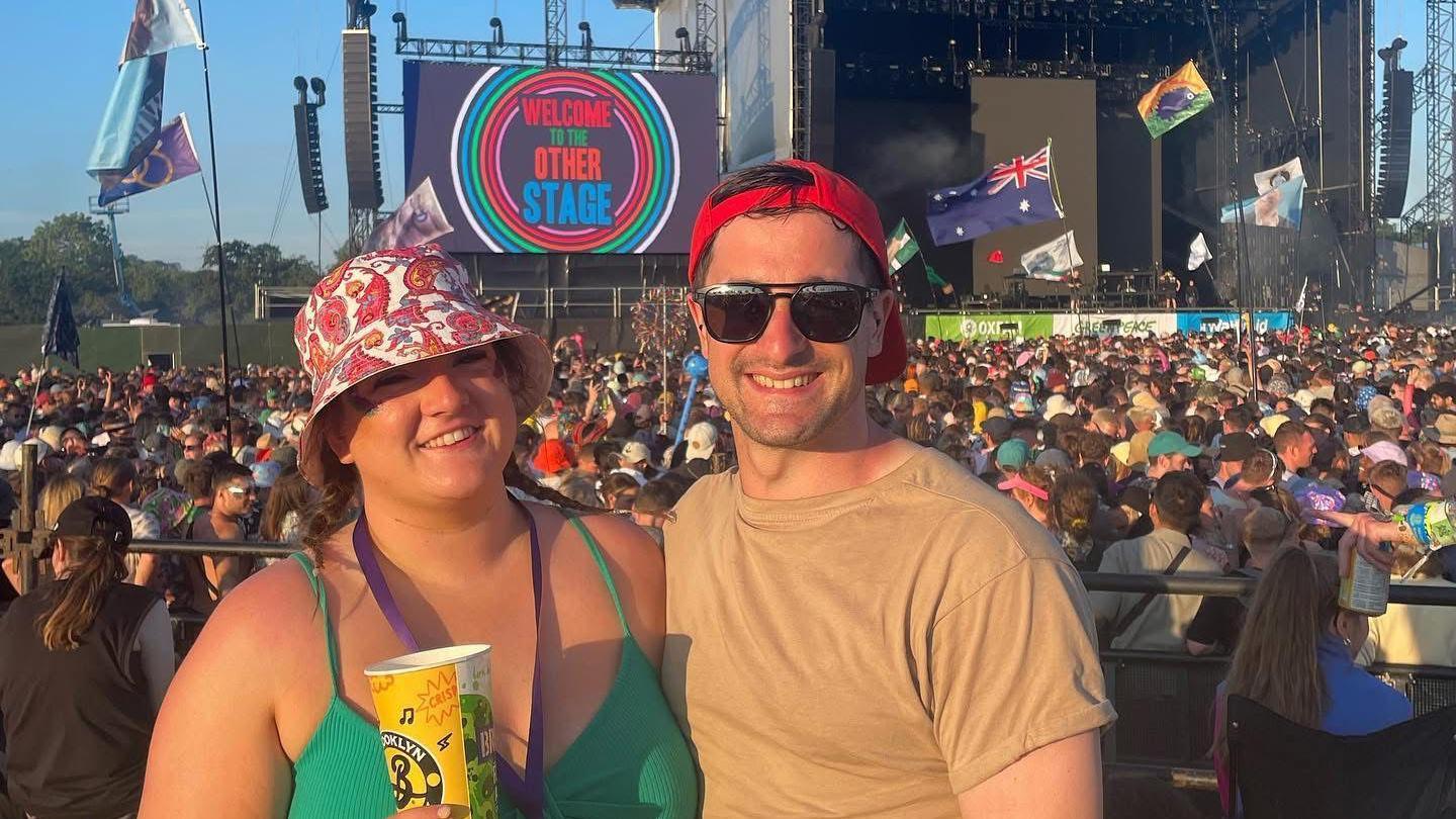 A woman and a man stand illuminated by sun in front of a festival stage, with a large crowd watching the stage.