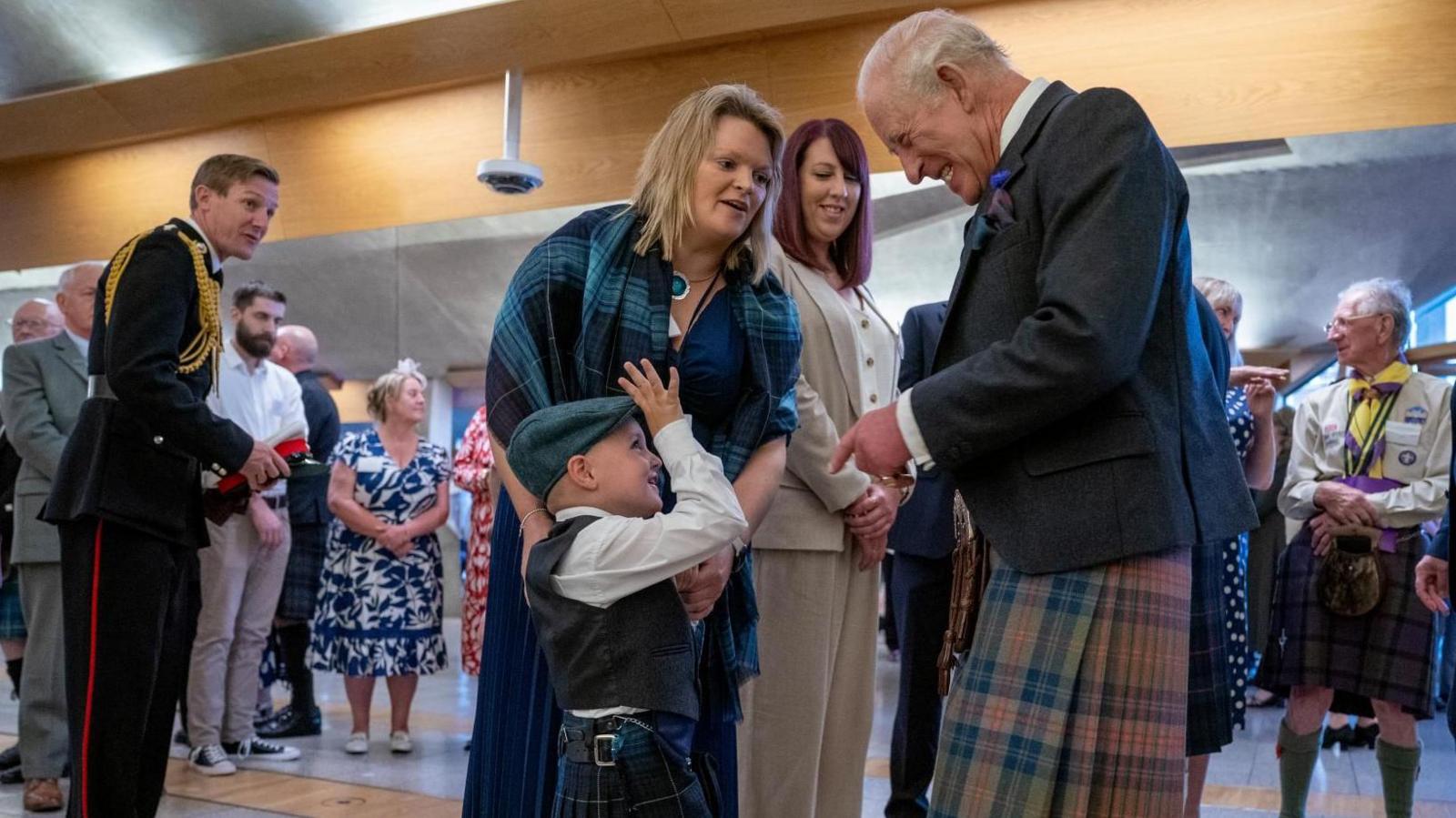 Four-year-old Gregor Corse holds up his his hand to King Charles with his fingers raised. They are both wearing traditional Scottish outfits including kilts. Gregor is also wearing a flat cap. Also pictured is Gregor's mum who is wearing a blue and green tartan shawl.