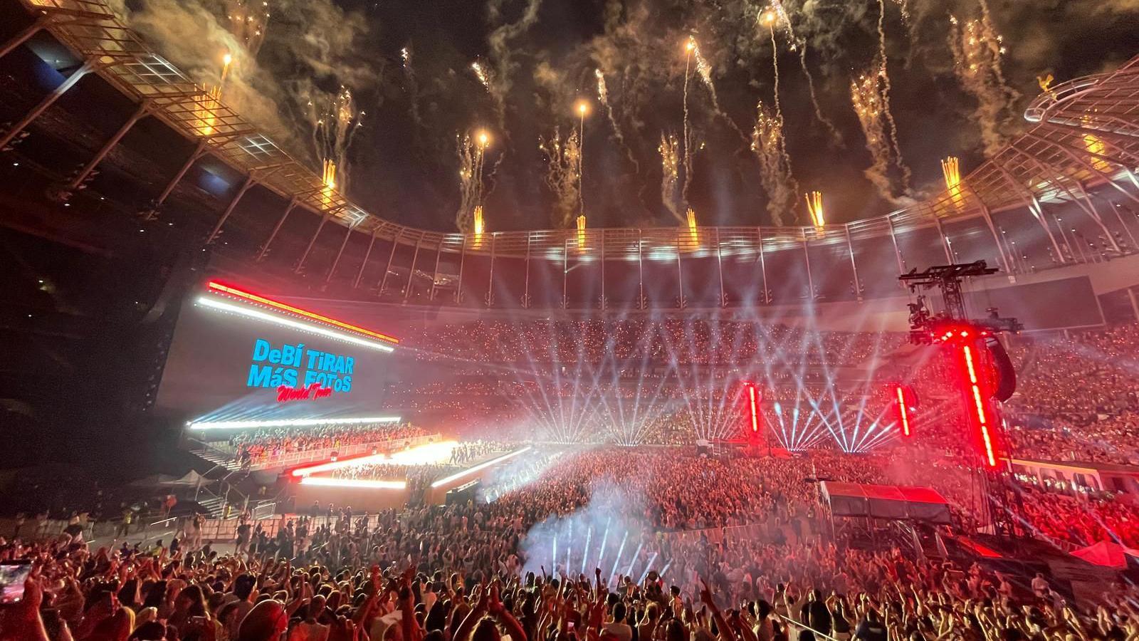 A huge crowd fills the Tottenham Hotspur Stadium, illuminated by stage lights. Fireworks go off from the roof. A stage is lit brightly. Behind, the big screen says Debí Tirar Más Fotos World Tour.