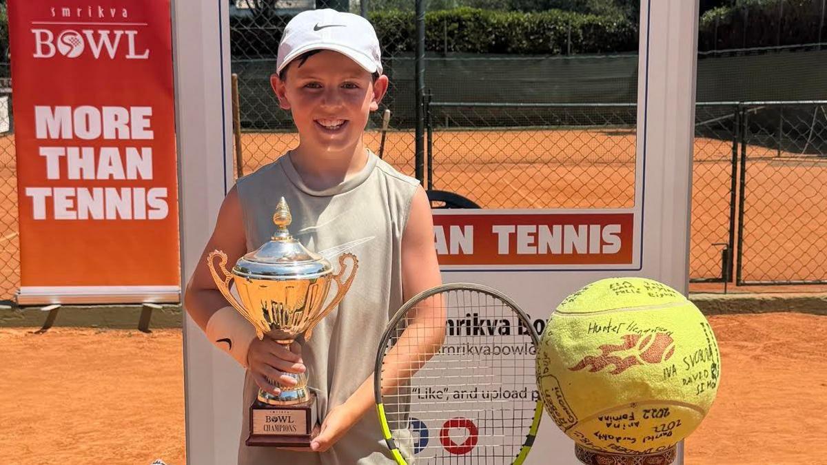 A young tennis player stands on a clay court holding a trophy, with a racket, bag, and signed tennis ball displayed nearby.  Behind the player, banners read “BOWL” and “MORE THAN TENNIS,” placed in front of a fenced court.  The scene is brightly lit under the sun, with organised items in the foreground suggesting a recent tournament win.