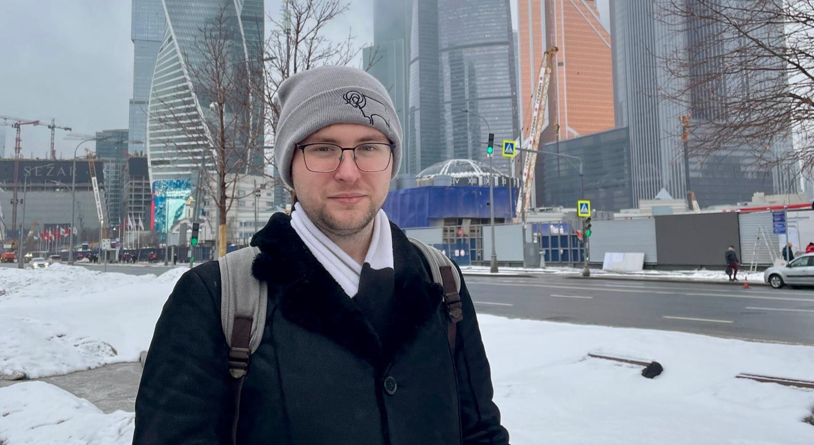 Person wearing a grey beanie, glasses, a black coat and a white scarf, carrying a grey backpack with visible shoulder straps in the foreground. Snow covers the pavement and roadside. A wide street with traffic lights and pedestrian crossing signs runs behind. In the background, modern high-rise buildings with glass façades and construction cranes are visible. Bare trees line the street, and temporary site structures and barriers are present near the road.