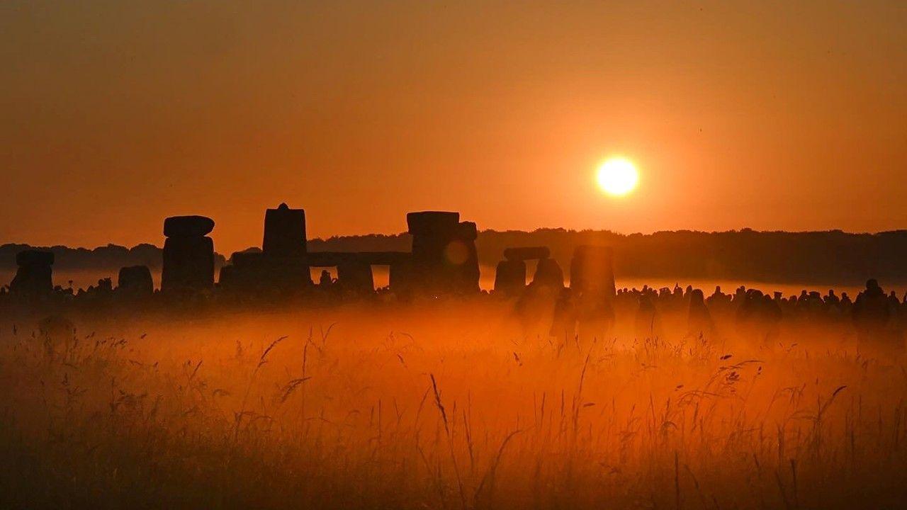 The Sun rising over the stones of the Stonehenge monument. The stone along with some people are silhouetted against the orange-tinted sky with the Sun to the right. The foreground is misty.