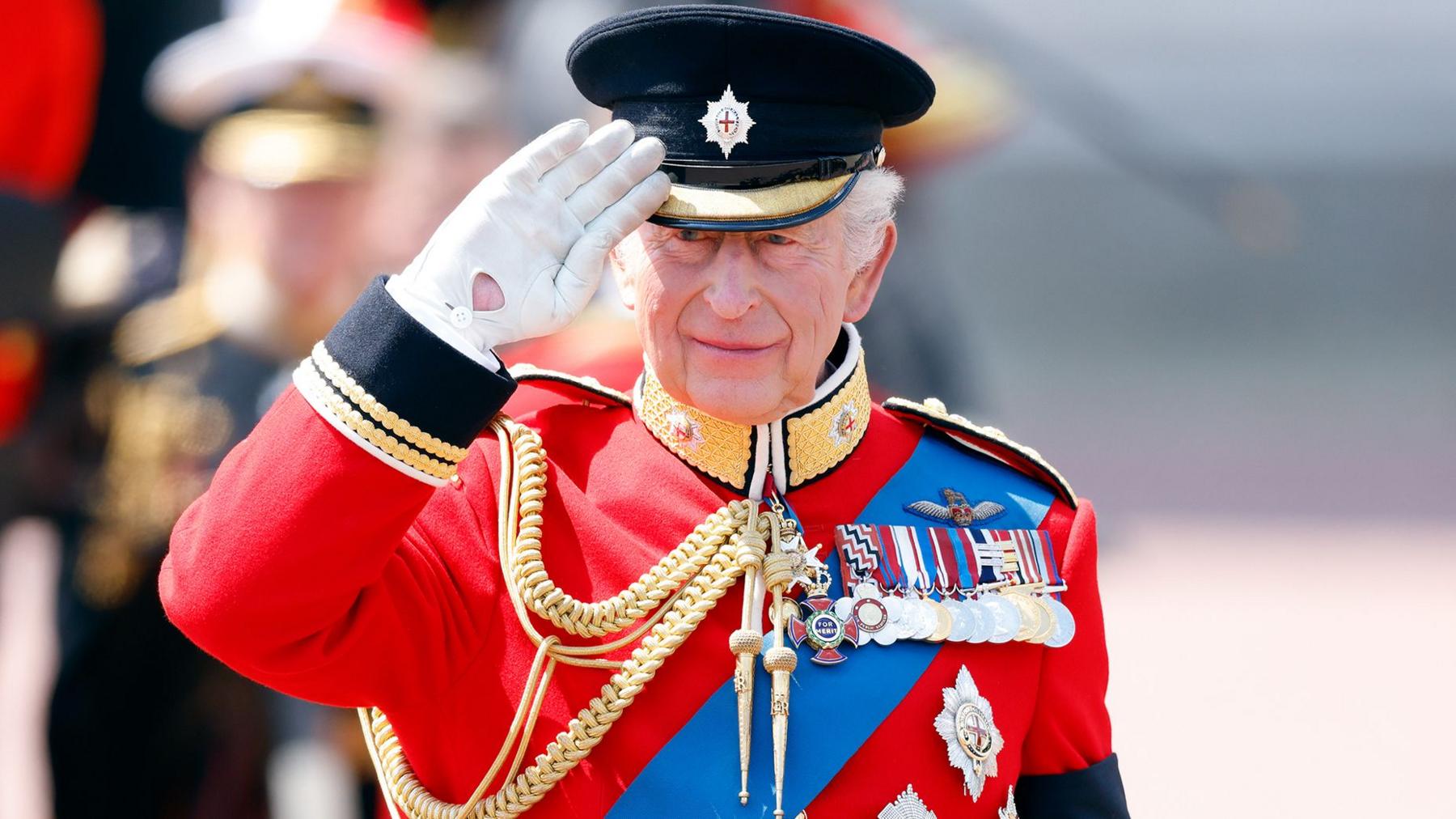 King Charles in a red military jacket and medals saluting to the left of the image   at the trooping of the colour ceremony in June 2026