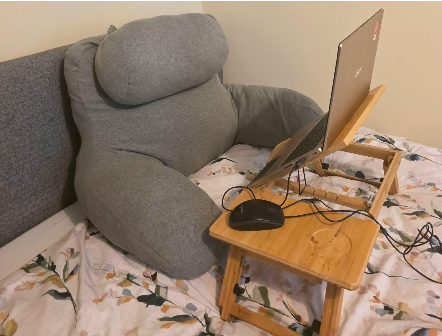A large support cushion leans against a grey headboard and a laptop sits on a small desk on the bed.