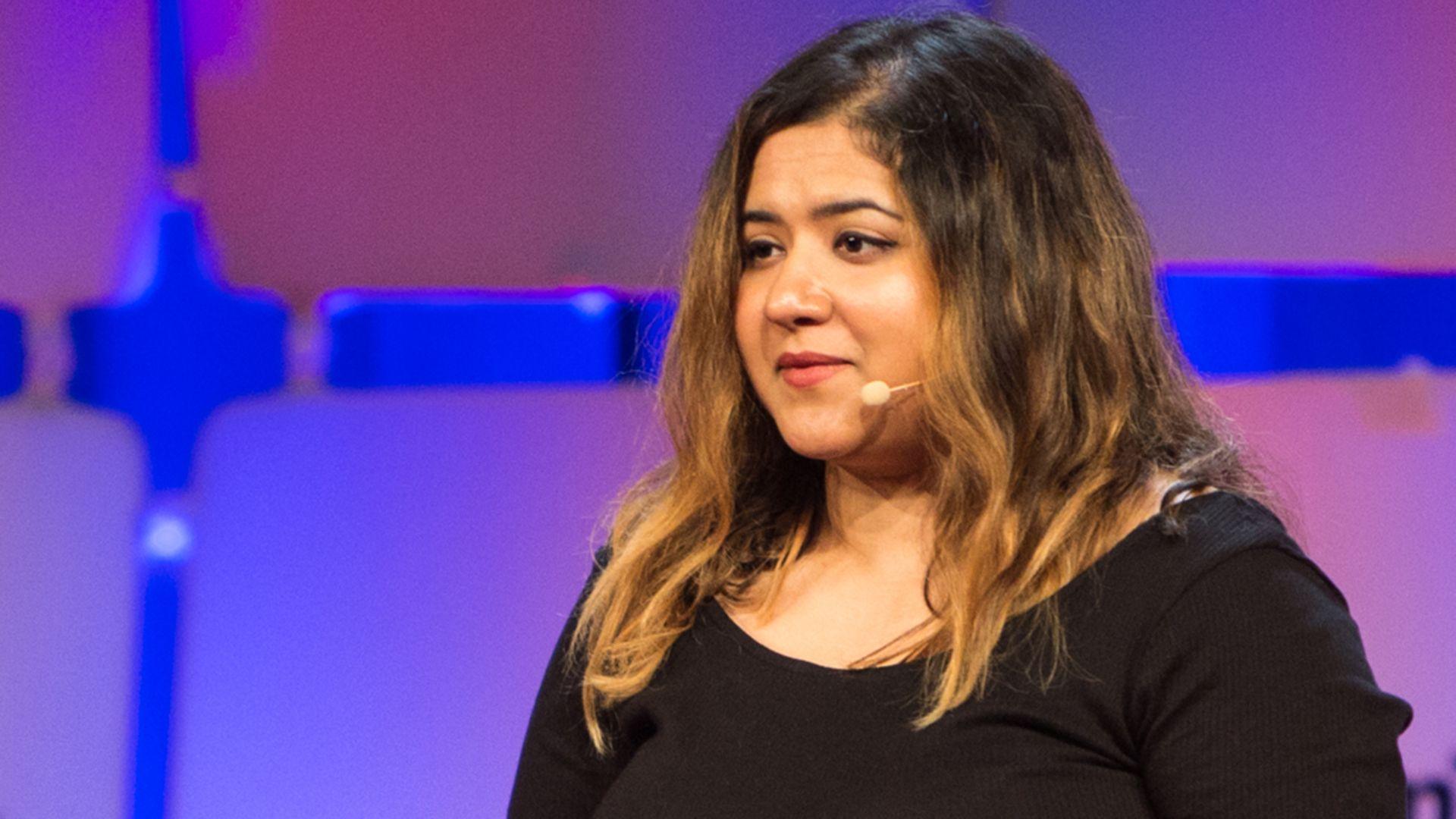 Headshot of a woman with long brown hair speaking at a conference. She is wearing a black top with long sleeves. She is wearing a head microphone.