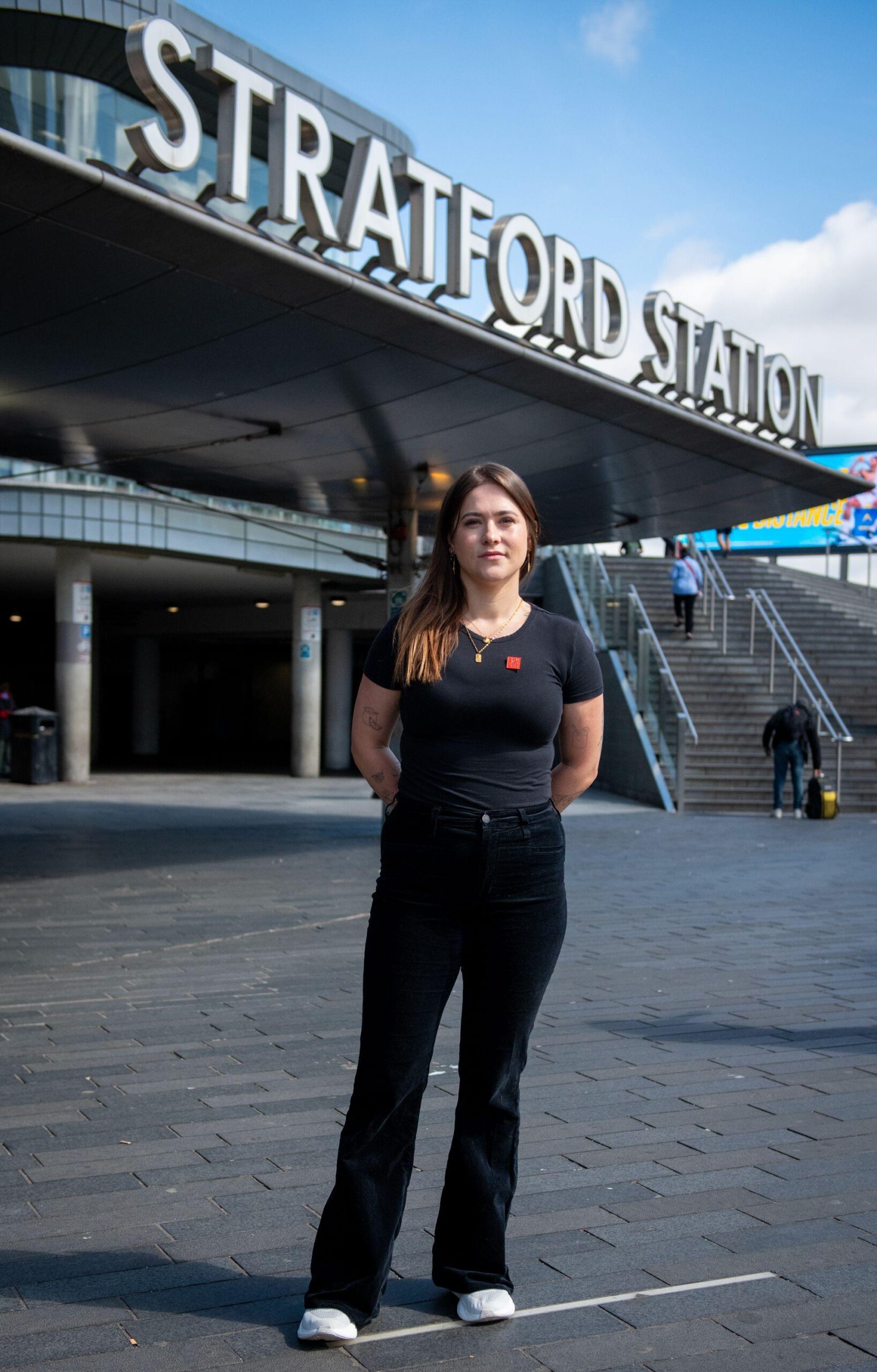 Esme Rice standing outside the entrance to Stratford Station, east London. It is a full-body shot, she is dressed all in black, the sign which says 'Stratford Station' is behind her.