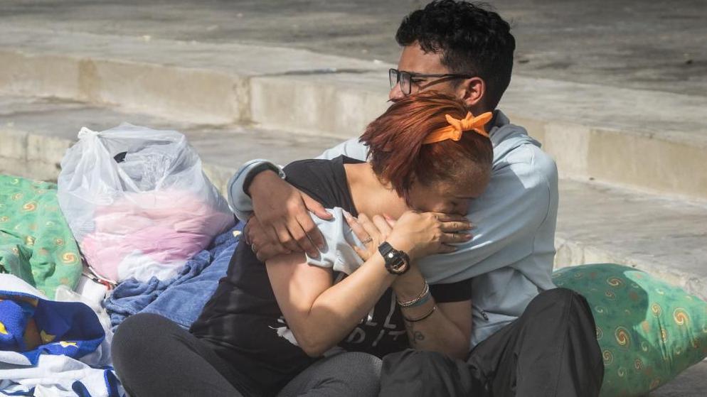 Two people cry into each others' arms surrounded by pillows, blankets and carrier bags of possessions on concrete stairs outside