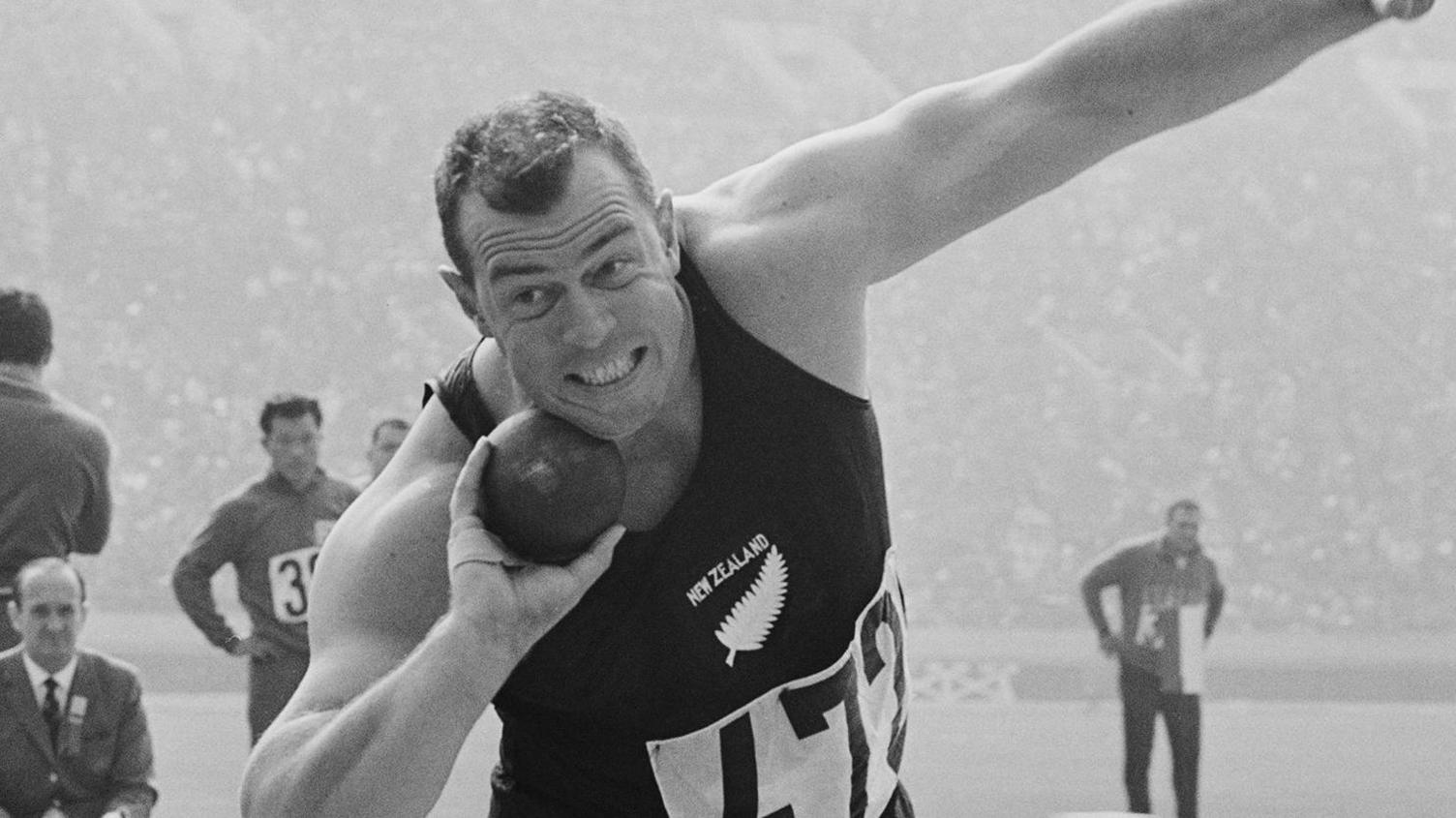 A black-and-white image of a shot-putter poised about to launch the putt - he has one arm holding the ball towards his face and the other arm out. He is wearing a black New Zealand bib with the number 472 - officials and a large stadium crowd are seen behind him