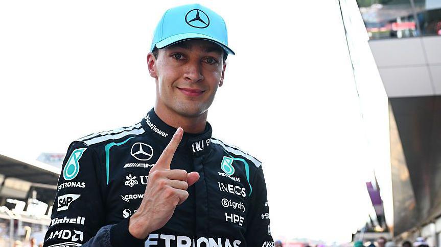 George Russell looks at the camera and points his index figure upwards