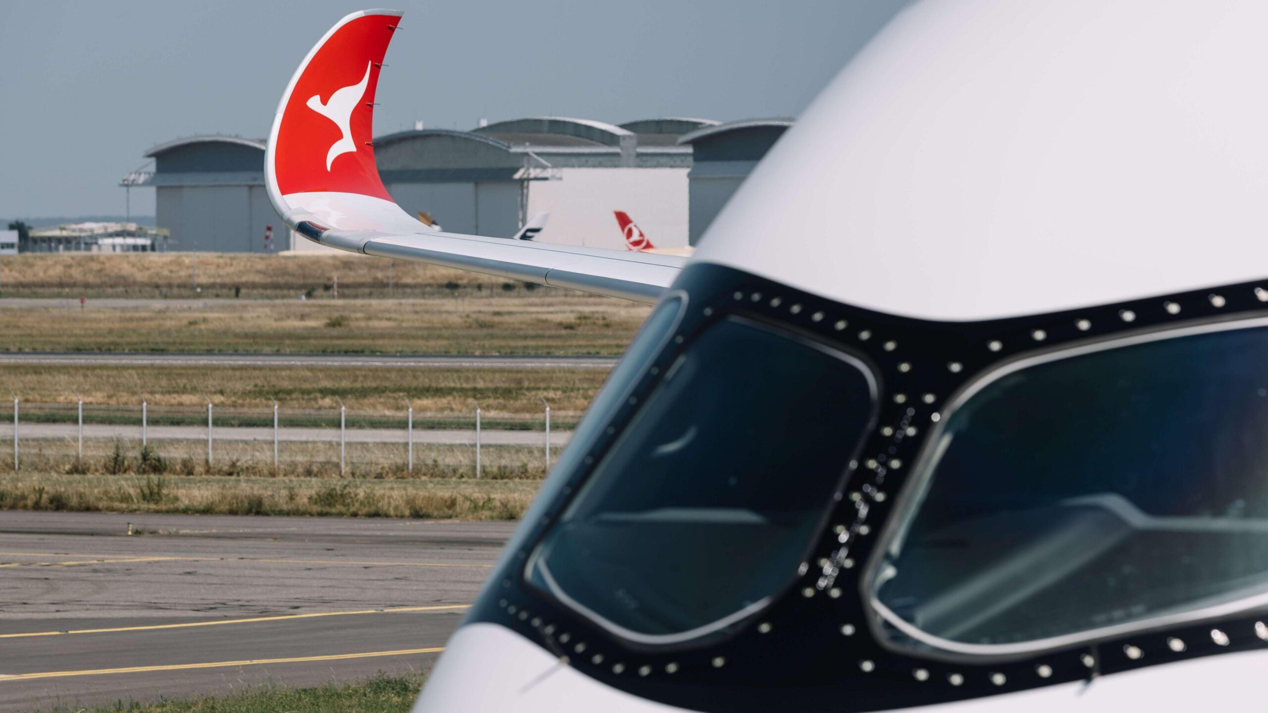 A close up of an airplane's cockpit, with the wingtip visible in the distance with the red and white kangaroo logo of Qantas.
