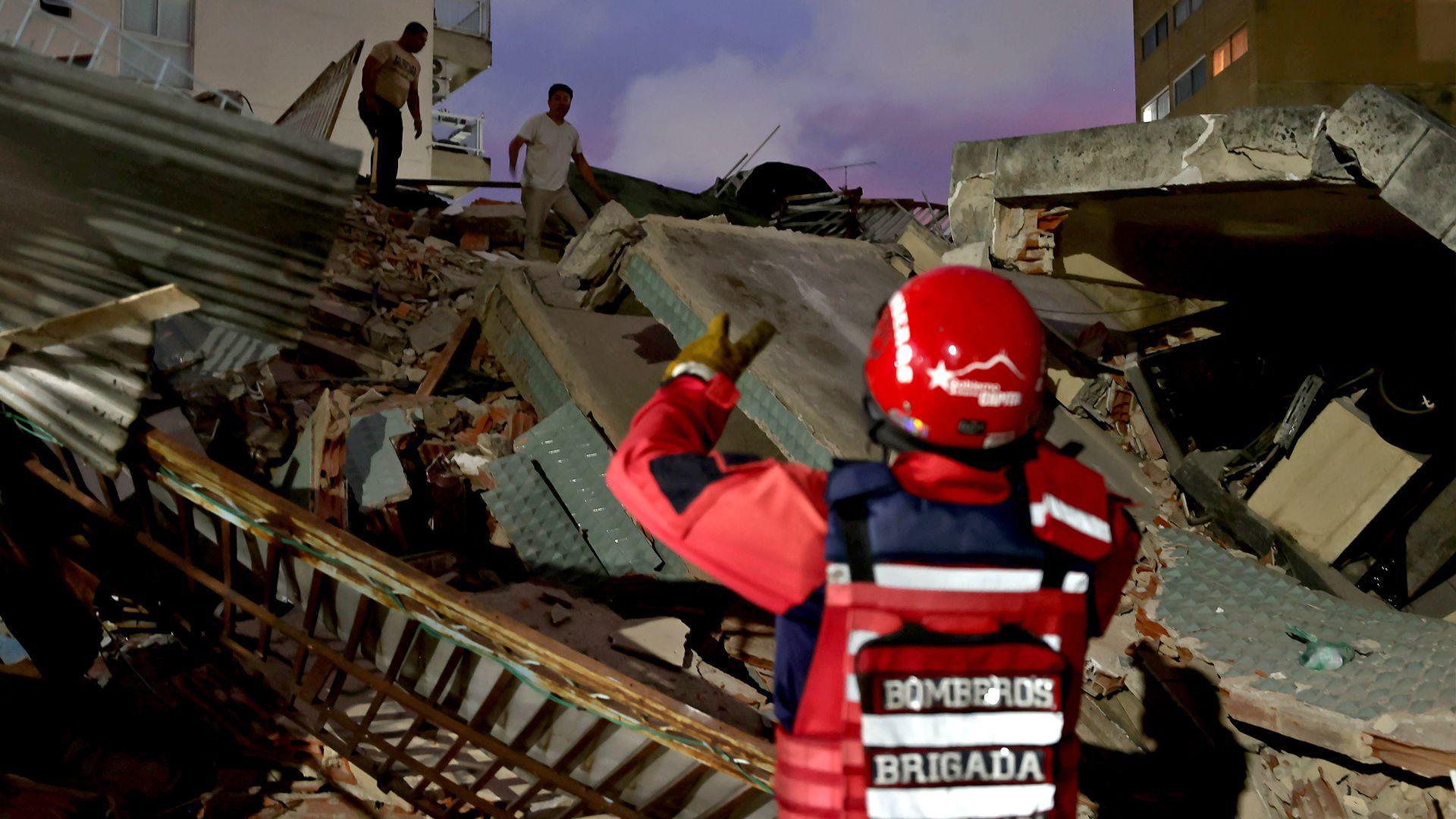 An emergency responder in red uniform including a protective jacket, helmet and vest, stands at the foot of a large pile of rubble and debris