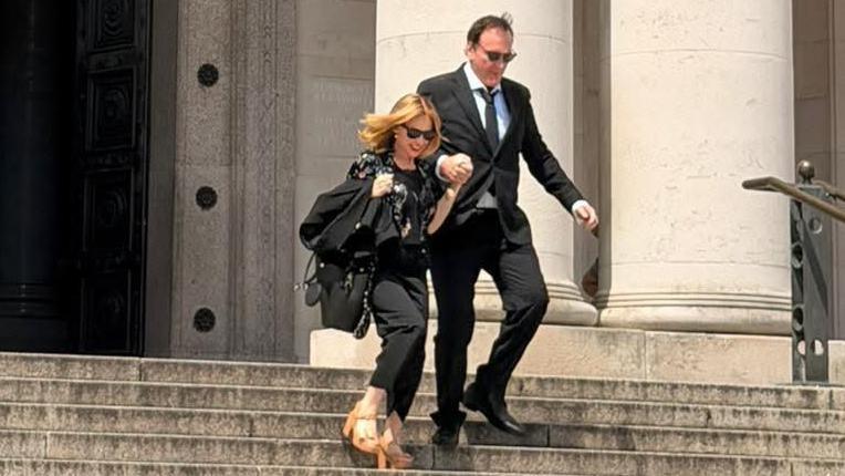 Quentin Tarantino and Kylie Minogue outside National Museum Cardiff. They are walking down the steps outside the museum holding hands. They are both wearing all black outfits and sunglasses. Kylie has a pair of brown tan high heels on.