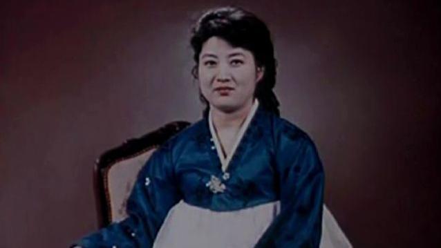 Ko Yong Hui, probably in her 30s or 40s, wears a blue and white Korean dress and smiles at the camera