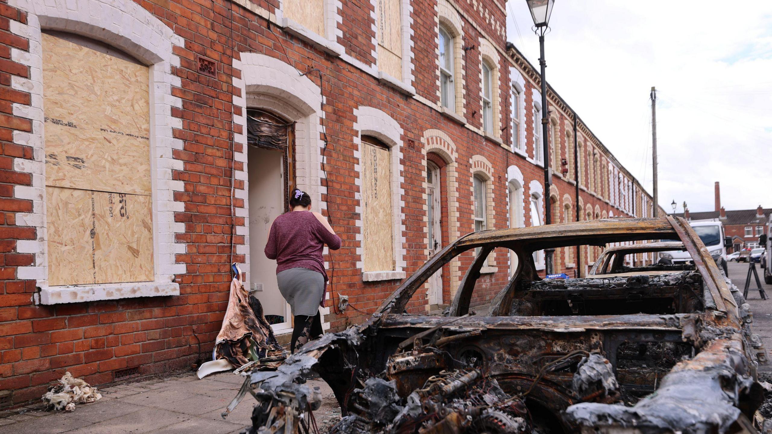 A burned-out car sits outside a row of boarded-up brick houses as a person walks into a damaged home.