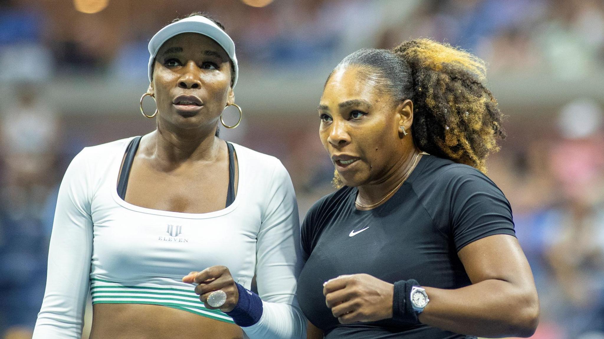 Venus Williams, in short white top and cap, stands alongside Serena Williams, who wears a black outfit