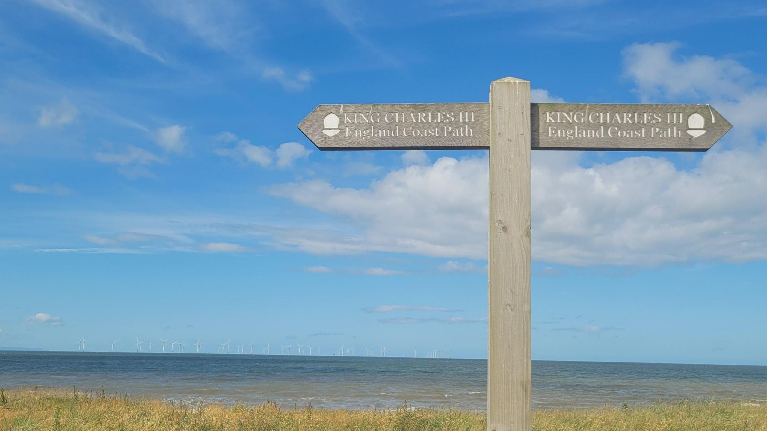 A fingerpost saying "King Charles II England Coastal Path", with the sea behind and a fairweather sky above