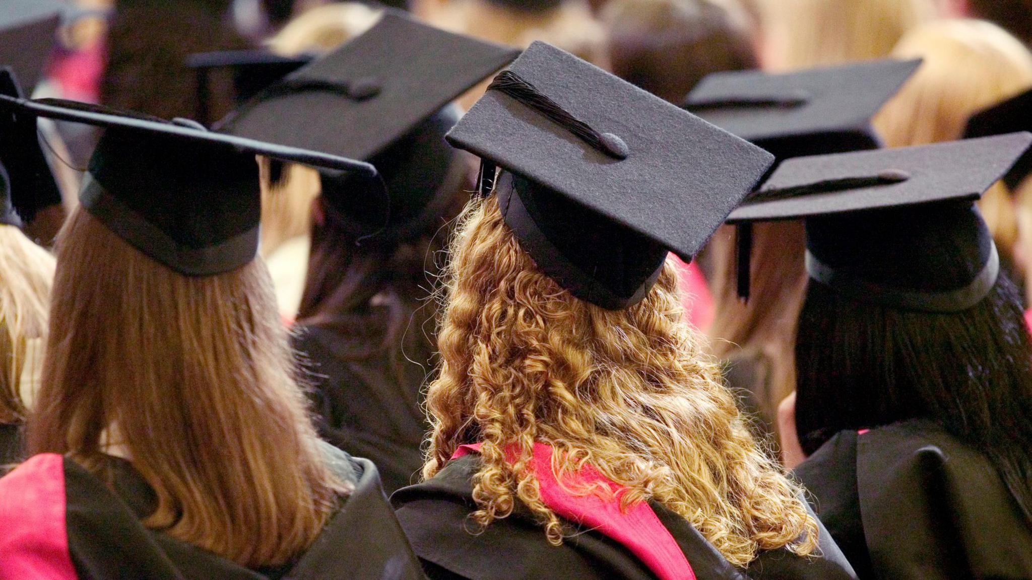 The image shows a graduation ceremony viewed from behind the audience of graduates. Several people are sitting closely together, all wearing black graduation gowns and square black caps with tassels hanging from them. One graduate has long, curly blonde hair spilling down the back of their gown, with a bright red sash draped over their shoulders.