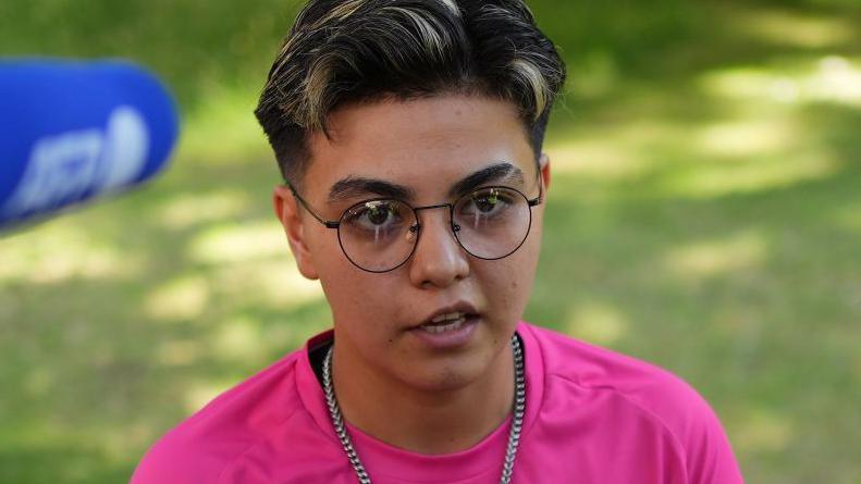 Ekil Latifi, a woman with cropped dark hair with highlights, wearing black wire-framed glasses and a silver chain necklace with a bright pink T-shirt