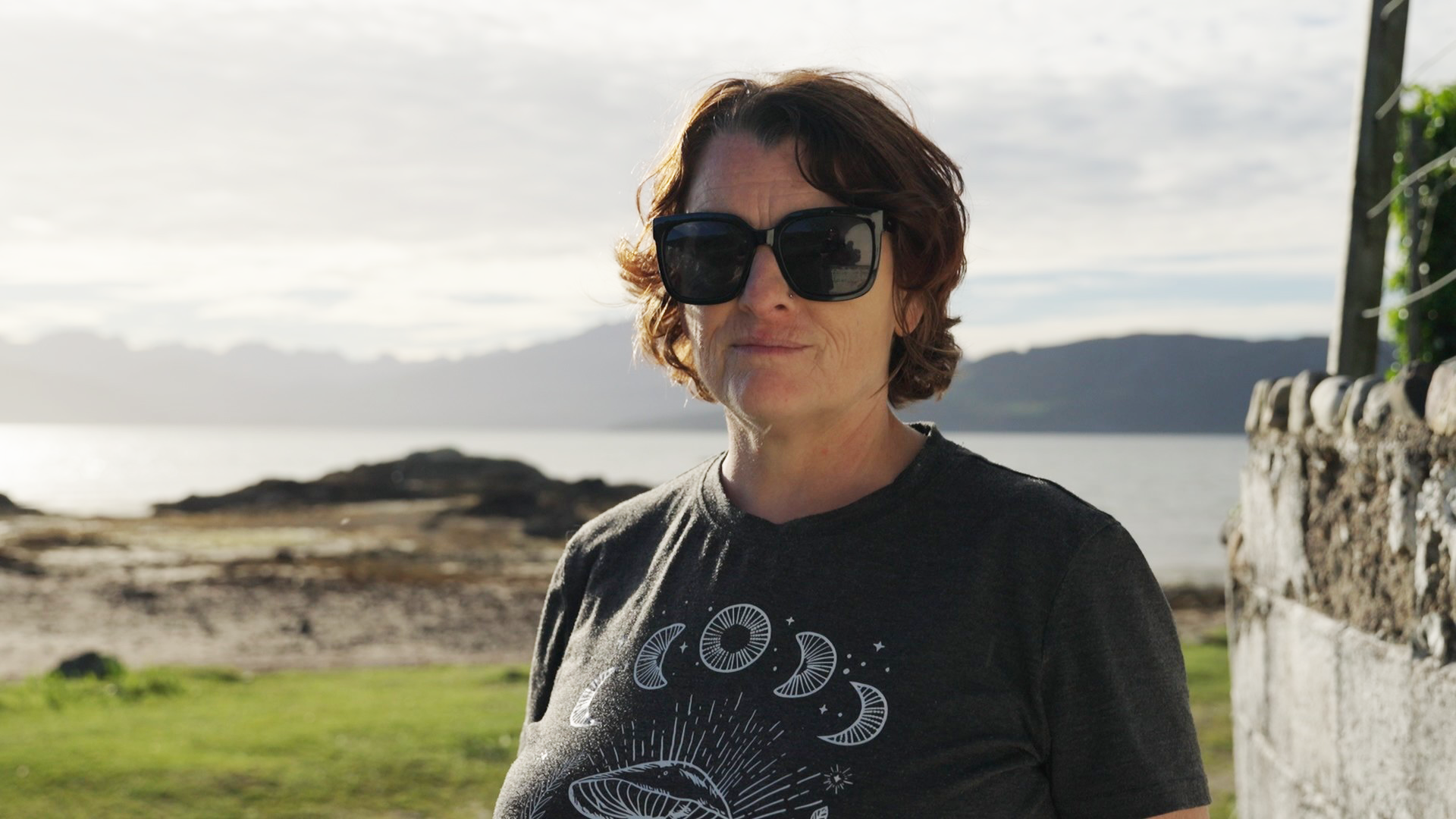 Julie Dawber has short, brown hair and is wearing a grey T-shirt and large sunglasses. She is standing next to a wall, with a view of Skye's coastline behind her.