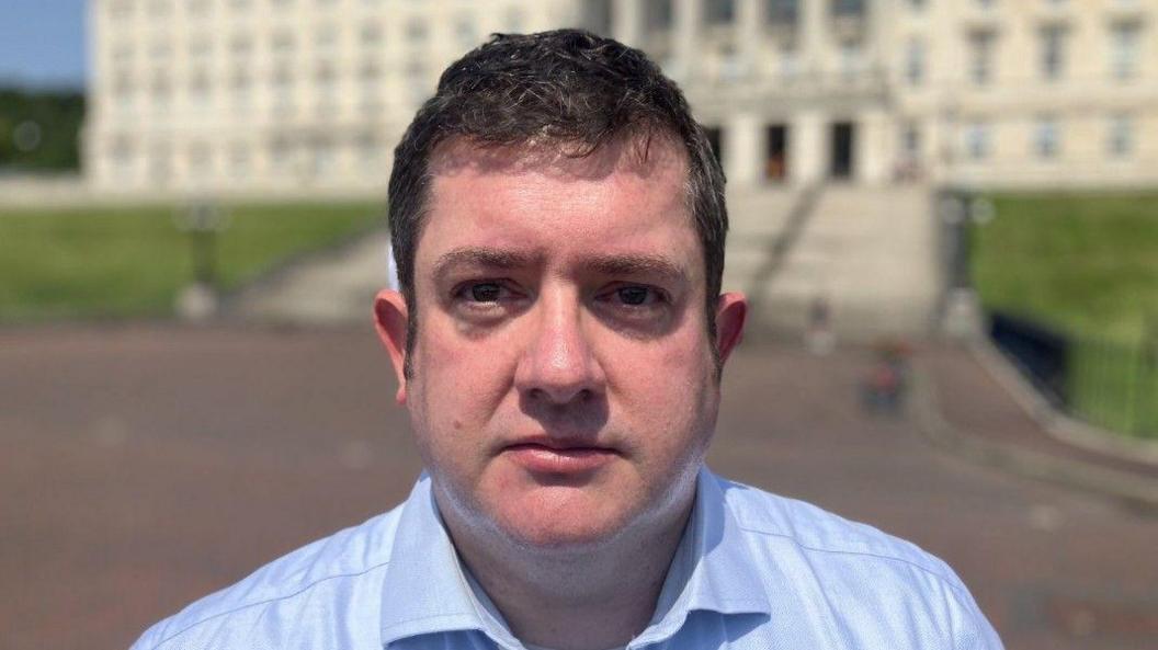 Dr David Farren, a man with dark hair, wearing a blue shirt, standing in front of Stormont Parlaiment Buildings.