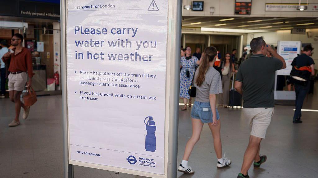 commuters walking past a transport for London sign saying 'please carry water with you in hot weather'