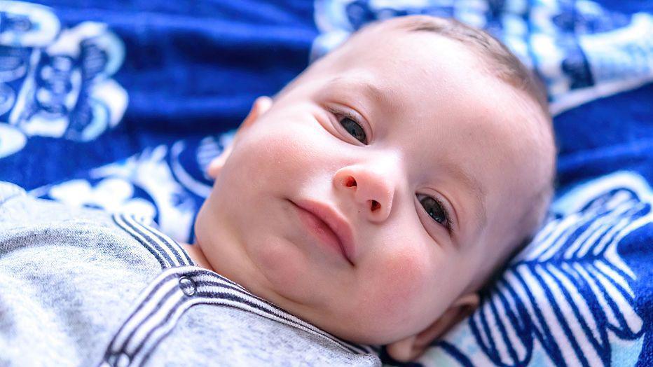 Medium shot portrait of baby boy. He is lying on a dark blue patterned fabric and wearing a grey sleepsuit
