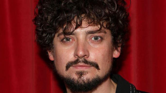 A man with curly dark hair and a dar beard and moustache. He is stood in front of a red curtain, wearing a glittery black shirt and looking at the camera with a serious expression.